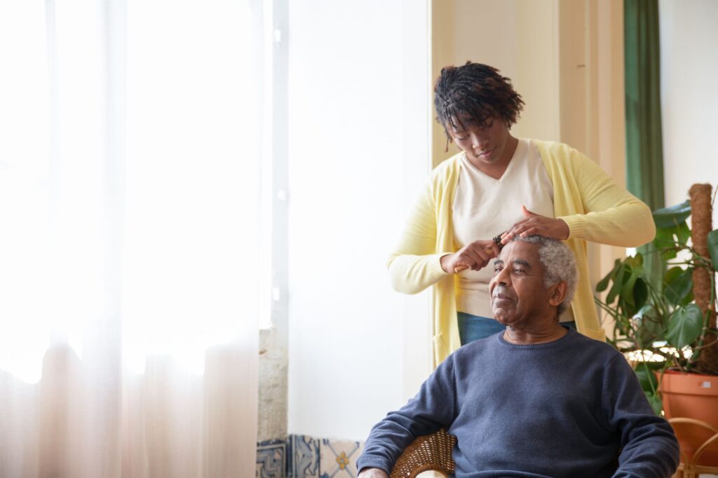 A caregiver combing an elderly man's hair near a sunlit window indoors.