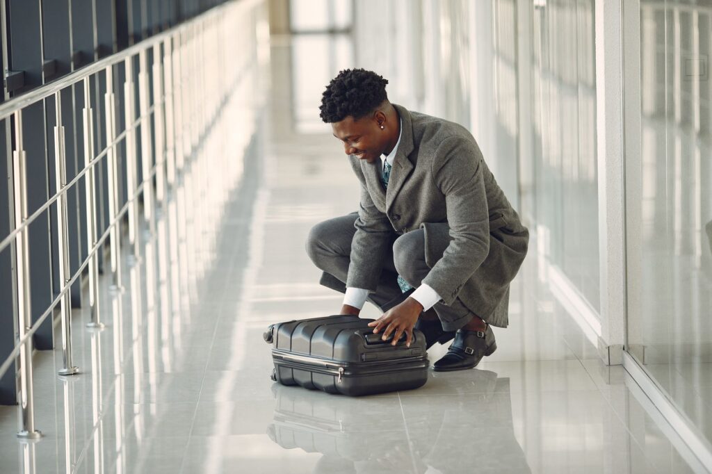 Full body of content black male passenger in classy gray clothes hunkering down to close suitcase while waiting for flight in modern airport hallway