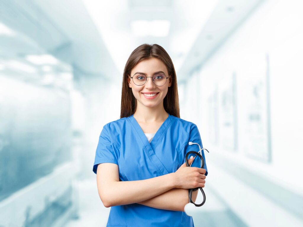 Young nurse in blue scrubs smiling and holding a stethoscope in a hospital corridor.