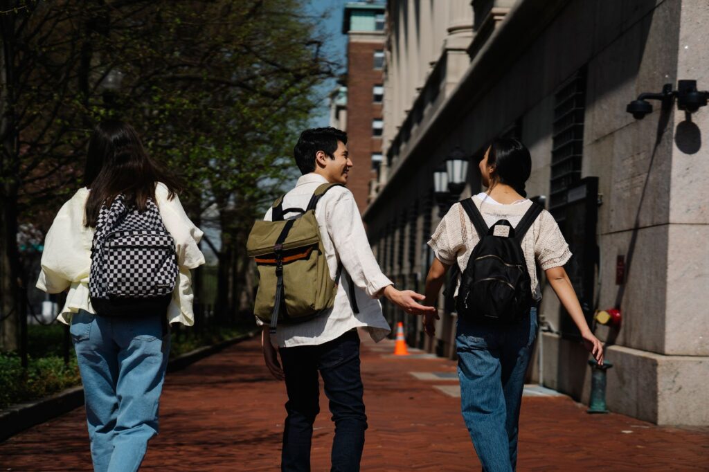 Three students with backpacks walk along a sunny campus path, engaged in friendly conversation.