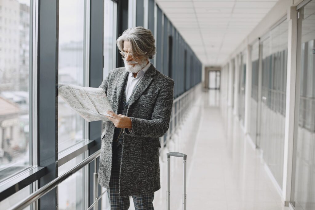 Senior man with beard reads map by glass windows in modern hallway with luggage.