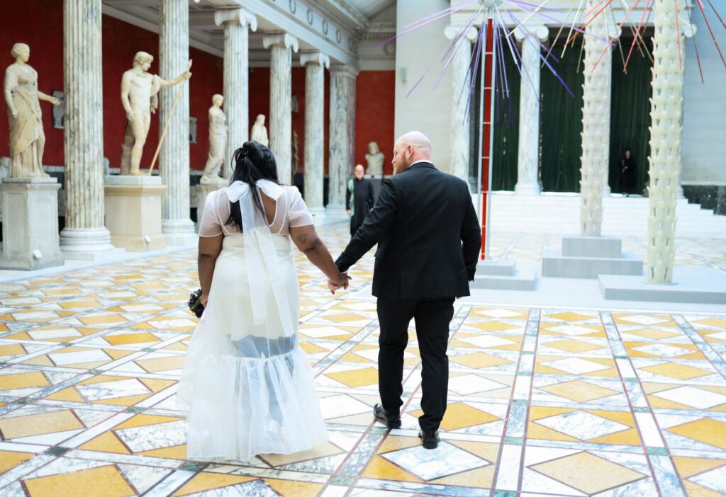 A couple holding hands in an ornate hall with classical statues and geometric flooring.