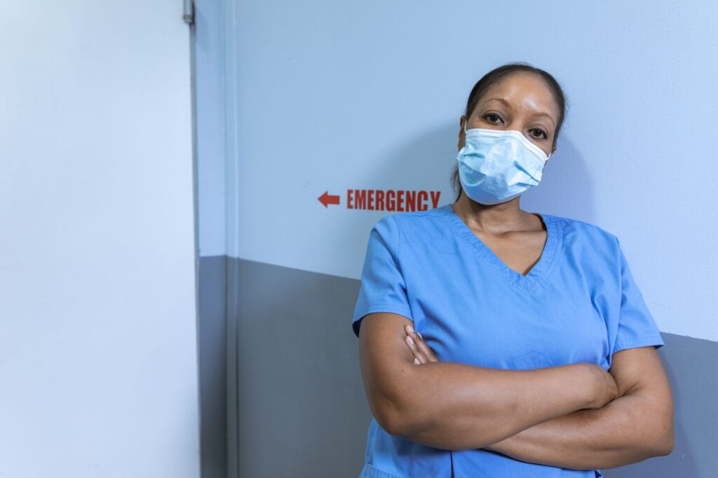 Nurse in blue scrubs and face mask in hospital corridor with emergency sign.