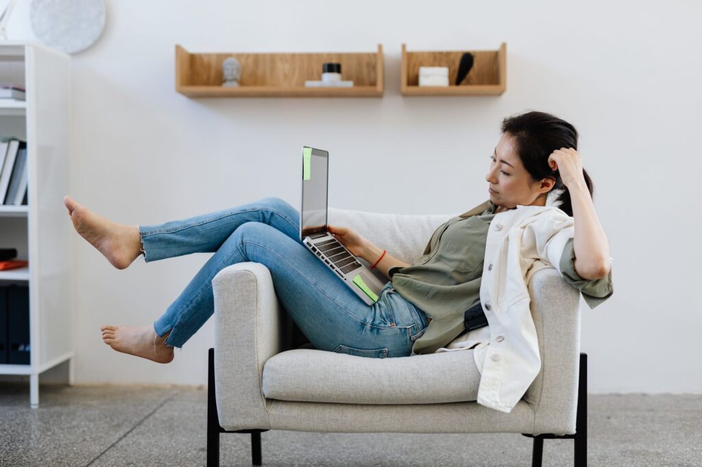 Asian woman lounging in an armchair, working remotely on a laptop at home.